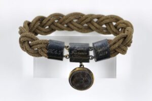 A woven bracelet, with a metal clasp and pendent that contains an lock of hair.
