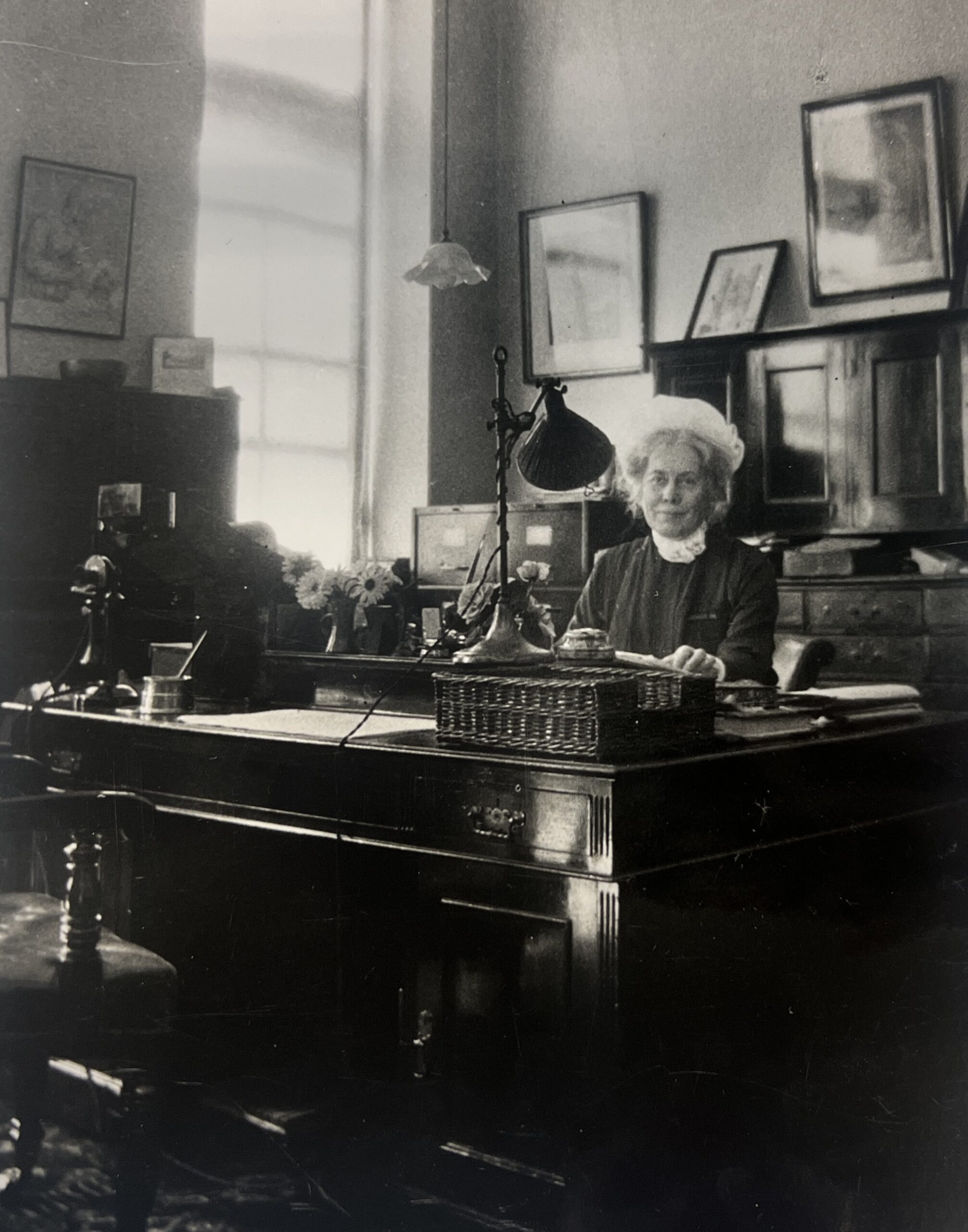 Black and white photograph of a lady sat at a large wooden desk. The room has dark paneling and framed pictures on the wall. The desk is very tidy and has a large lamp.