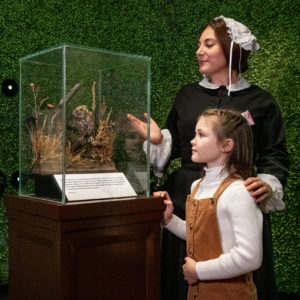 Florence Nightingale performer gestures towards a taxidermy owl in a glass case, standing next to a girl of about 10 years old