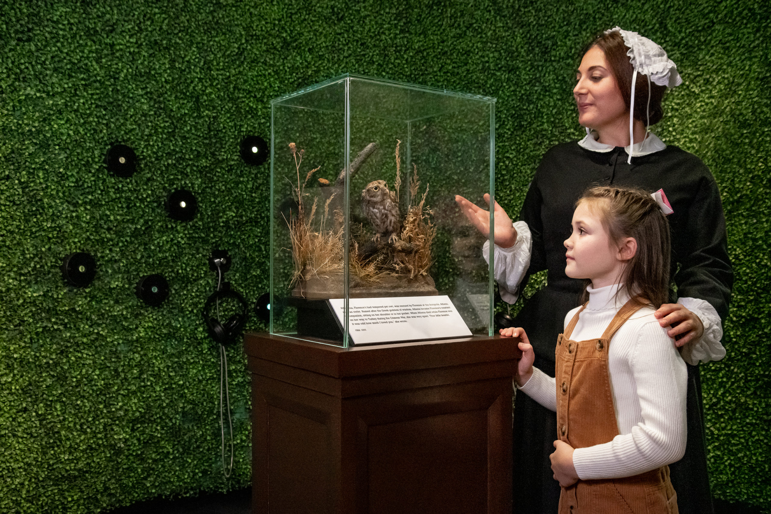 Florence Nightingale performer gestures towards a taxidermy owl in a glass case, standing next to a girl of about 10 years old