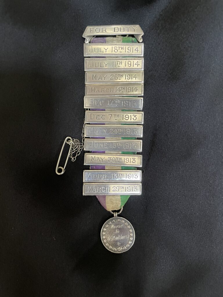A medal with silver coin at the bottom. The ribbon is green, white and purple, and contains 11 silver bars, all featuring dates.