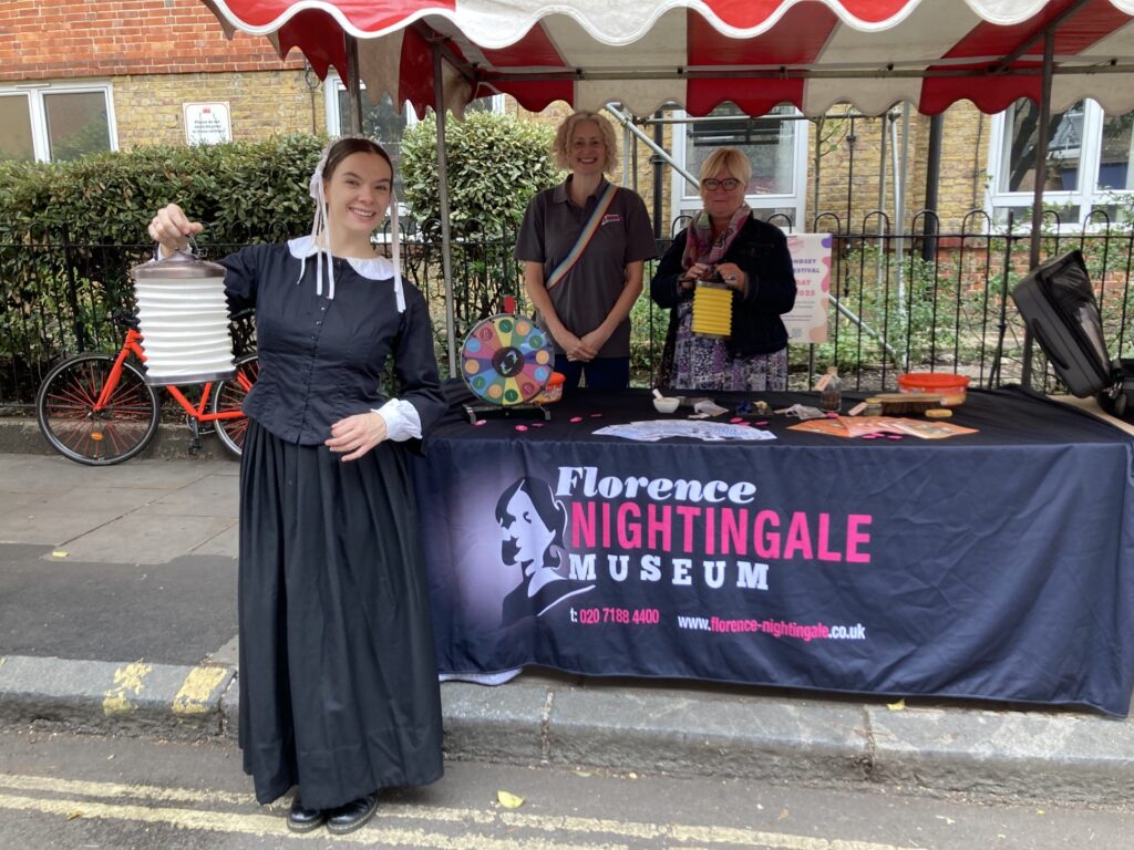 A Florence Nightingale actor stands in front of a marquee, 2 volunteers stand smiling behind