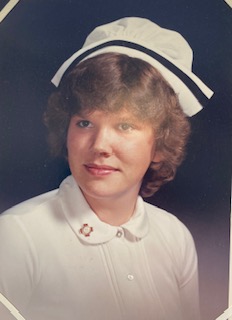 A graduate nurse wearing a nursing cap smiles softly in a formal portrait photograph. a red nursing pin can be seen on her lapel