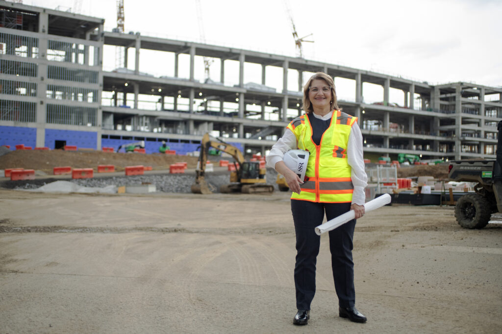 A lady wearing a hi-vis vest stands on a construction site, holding a hard hat and large rolled up piece of paper.