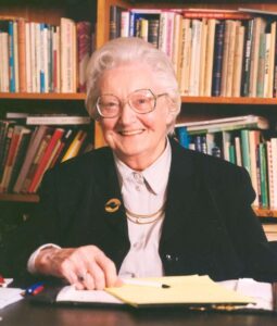Dame-Cicely-Saunders-reading-871×1024 an older lady with white hair and glasses smiles sat infront of a bookcase
