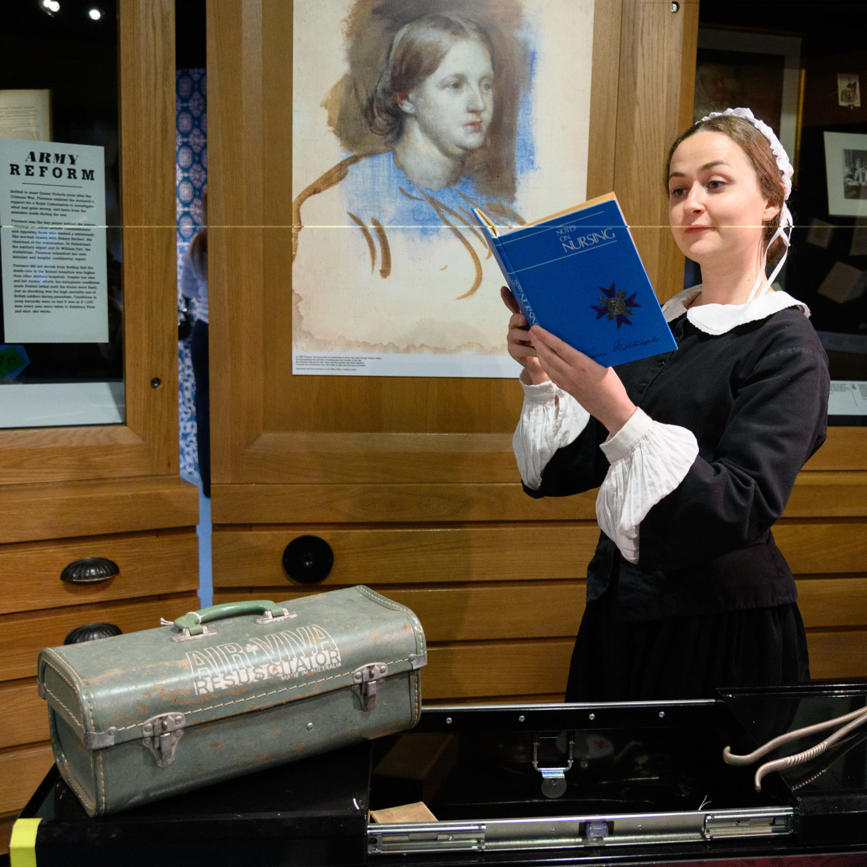 Florence Nightingale performer reads a copy of her book 'Notes on Nursing'. She stands behind a table displaying old medical equipment.
