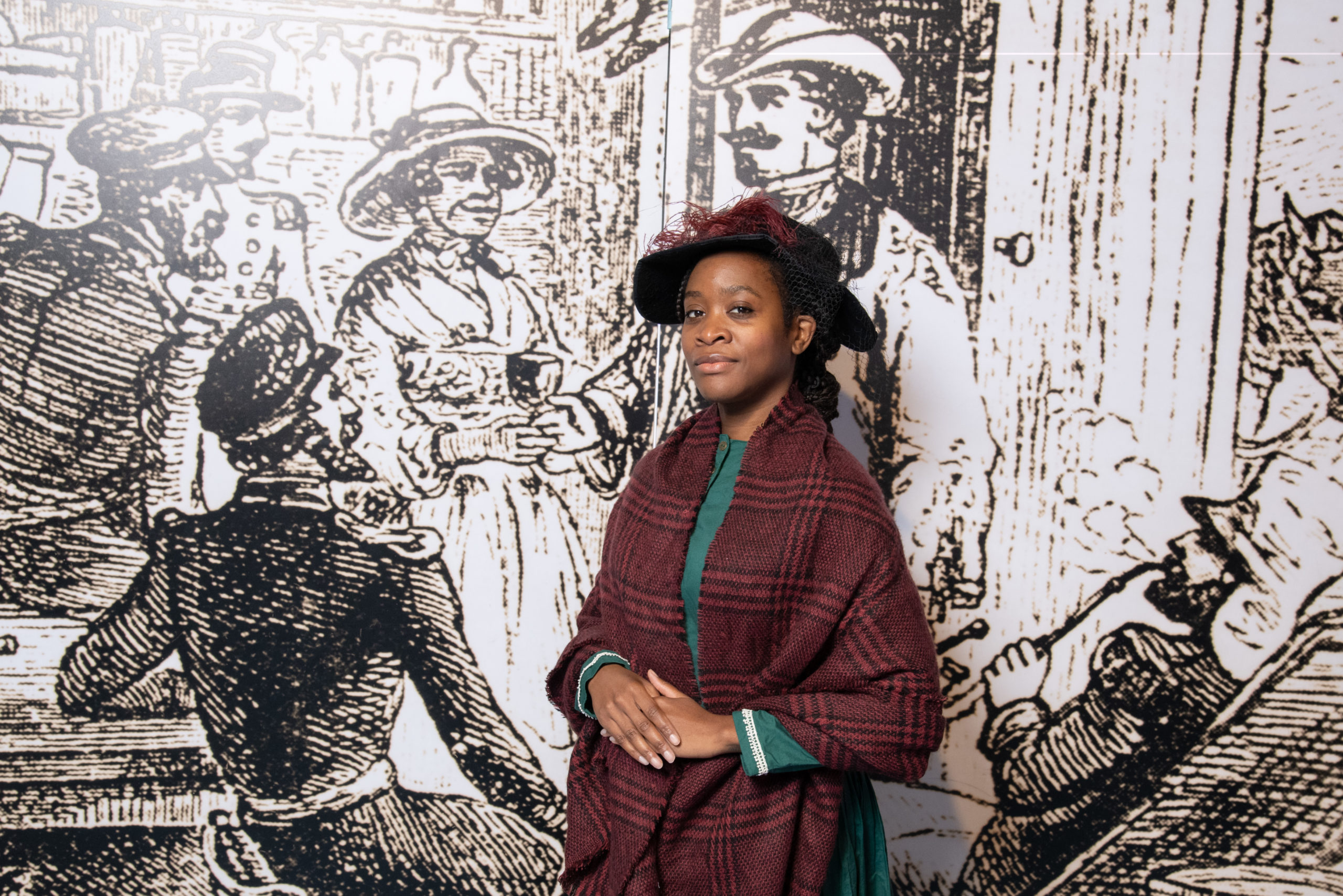 Mary Seacole performer poses for a photo. She wears a teal dress, maroon plaid shawl, and black felt hat. A maroon feather accents its wide brim.