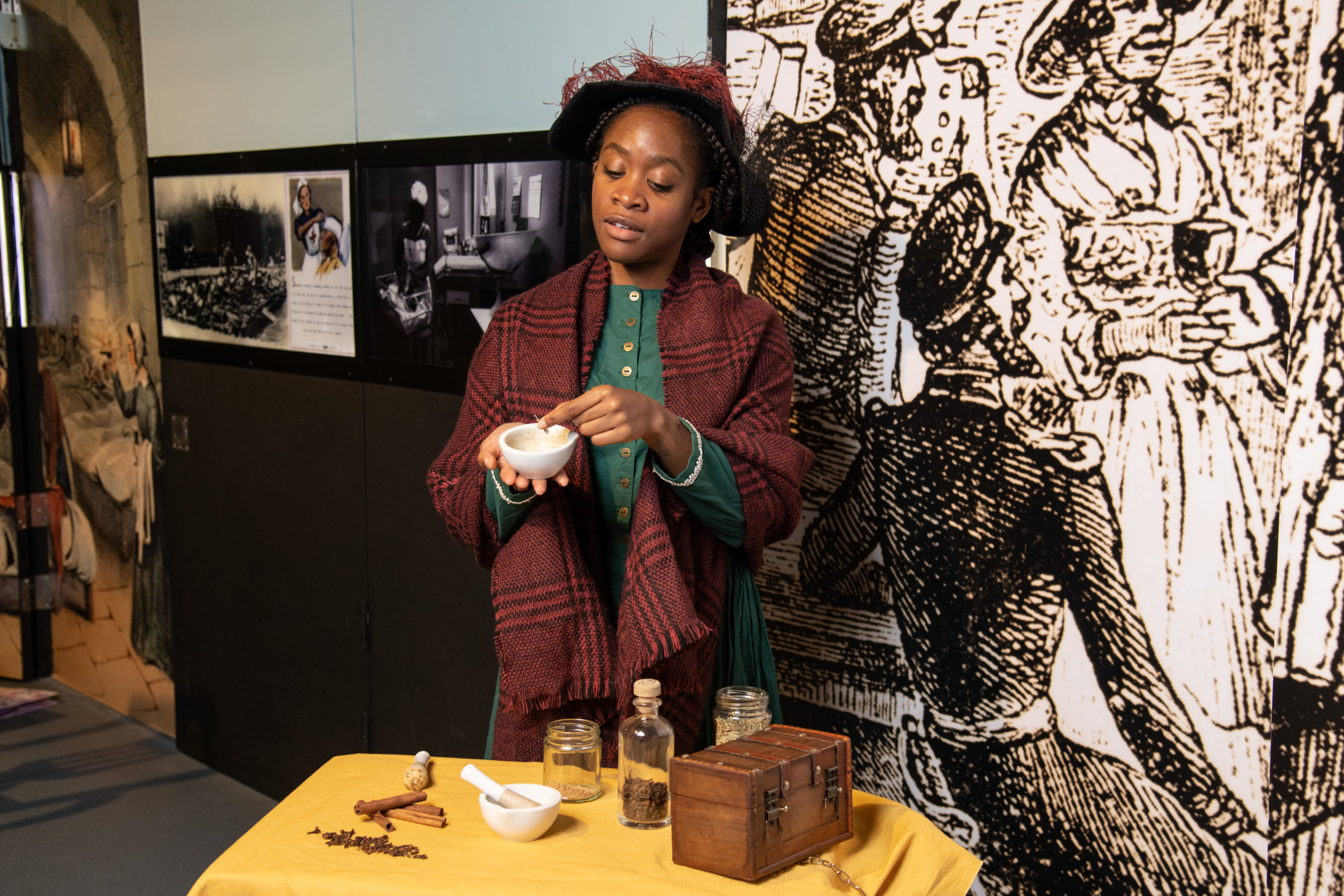 Mary Seacole performer poses for a photo. She wears a teal dress, maroon plaid shawl, and black felt hat. A maroon feather accents its wide brim.