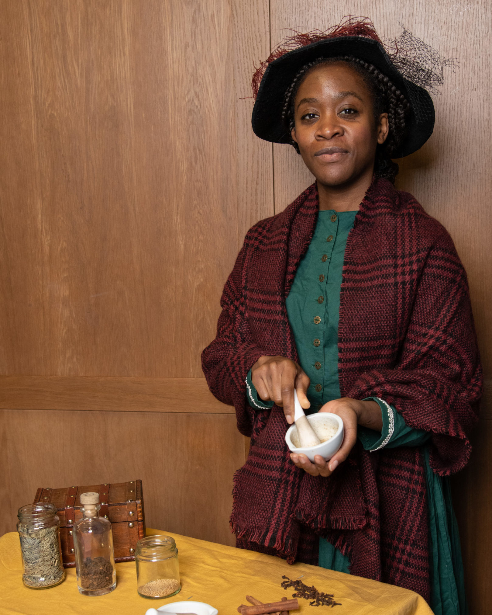 Mary Seacole performer poses for a photo. She wears a teal dress, maroon plaid shawl, and black felt hat. A maroon feather accents its wide brim.