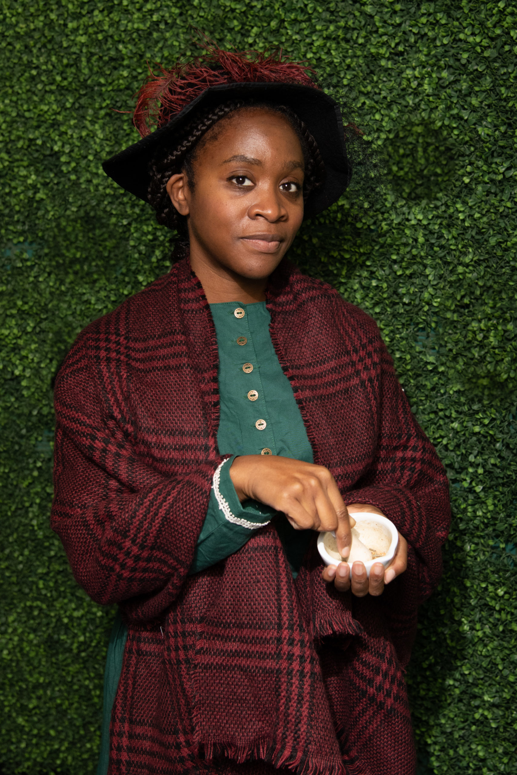 Mary Seacole photoshoot Mary Seacole performer poses for a photo. She wears a teal dress, maroon plaid shawl, and black felt hat. A maroon feather accents its wide brim.