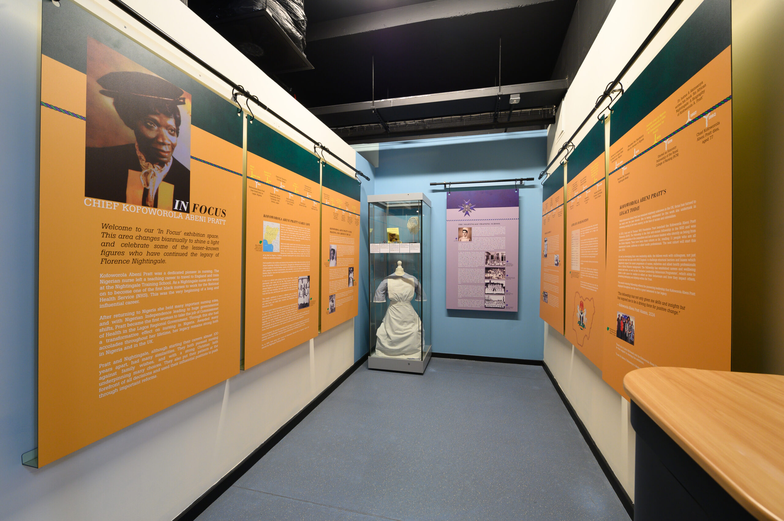 FNM_250127_095 A small room that has seven yellow panels hanging on the wall. This is the exhibition on Kofoworola Abeni Pratt. Each panel has text and images. At the end of the room is a display case that contains objects.