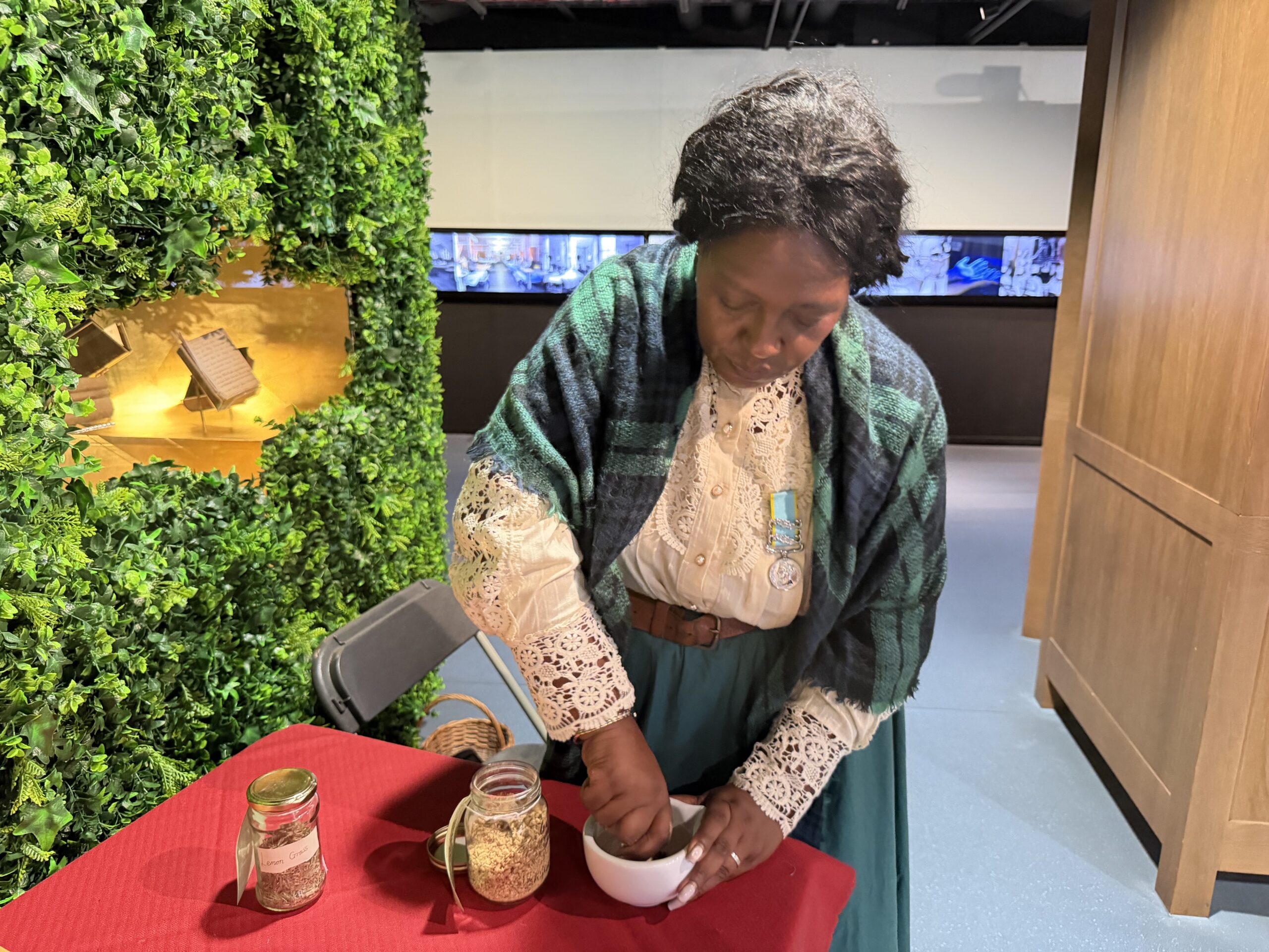 An actress dressed as Mary Seacole stands at a table with a red tablecloth using a pestle and mortar to mix together herbs. Two open jars are next to her, one labelled ginger and the other labelled lemon grass.