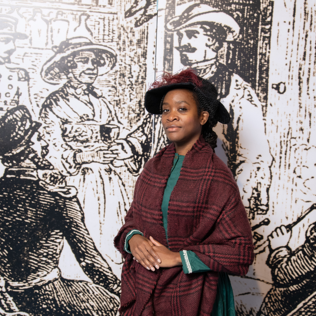 Mary Seacole performer poses for a photo. She wears a teal dress, maroon plaid shawl, and black felt hat. A maroon feather accents its wide brim.