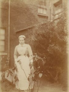 A sepia photograph of a lady in nursing uniform. She is stood in front of a brick building.
