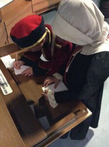 Photo 06-08-2023, 13 10 20 two children write letters on a wooden drawer. one is dressed in a red soldiers outfit and the other in a black nurses dress with bonnet