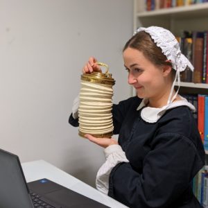 Florence Nightingale performer sits in front of a laptop. She raises a paper lantern level with the laptop's camera.