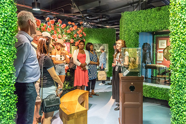 In the exhibition space, nine visitors stand viewing different displays. A number are looking at a tour guide, who is talking. There is a bench for public use and display cases containing artifacts.